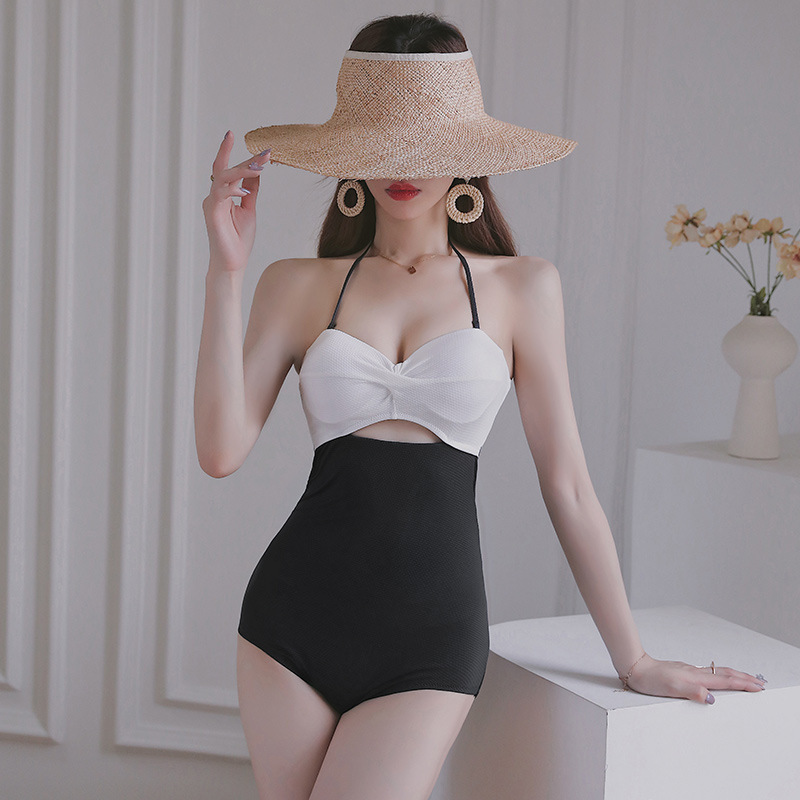 Front view of black and white two-tone halter one-piece swimsuit with gathered white bust and small front cut-out, model resting one hand on a plinth